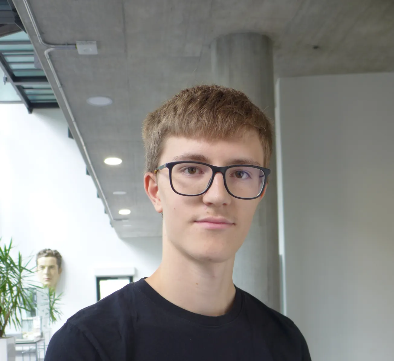 Close portrait of James Bennett, wearing dark-framed glasses and a black t-shirt, in a bright concrete and glass building