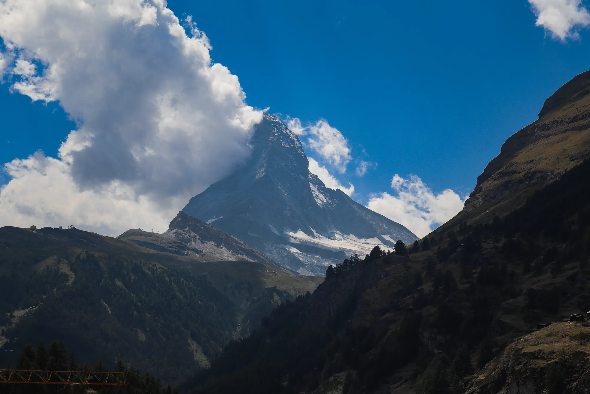 A rocky mountain peak with snow patches, partly wrapped in cloud, above a dark forested valley