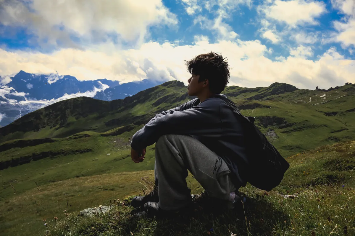 James sitting on a grassy ridge in profile, looking out over green slopes and distant snowy mountains