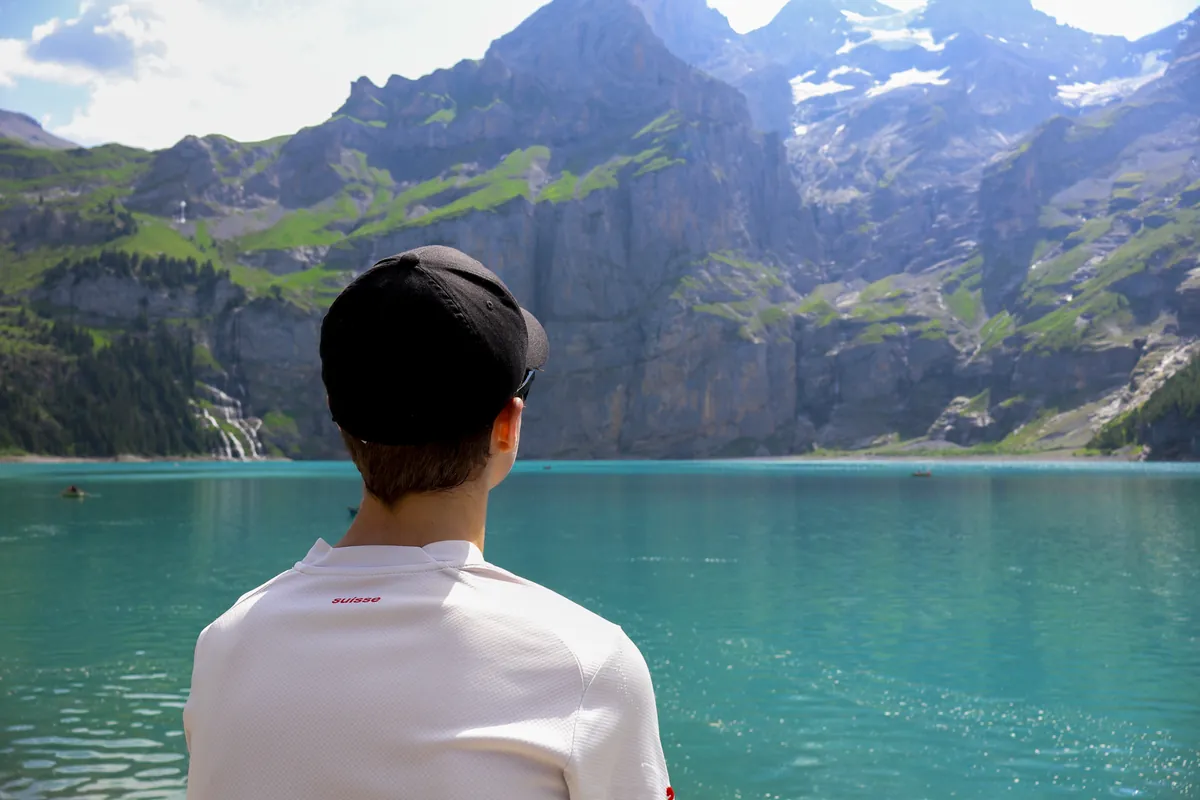 James seen from behind in a white shirt and black cap, looking across a turquoise mountain lake