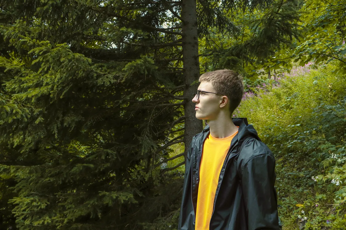 James in a dark jacket and yellow shirt, standing in a green forest and looking to the side