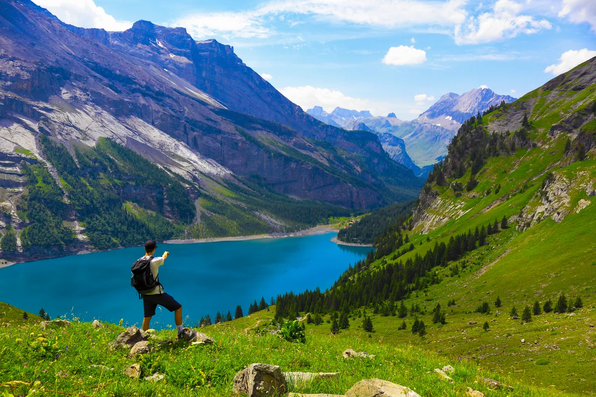 A hiker with a backpack standing on a grassy slope above a bright blue mountain lake